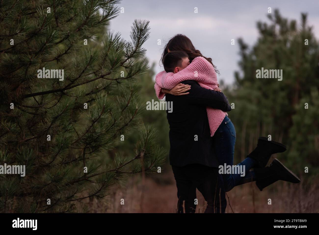 La coppia felice gira nella foresta di conifere lungo il sentiero. Il giovane abbraccia strettamente la donna dai capelli ricci con un maglione rosa. Fidanzamento invernale, matrimonio autunnale. Proposa Foto Stock