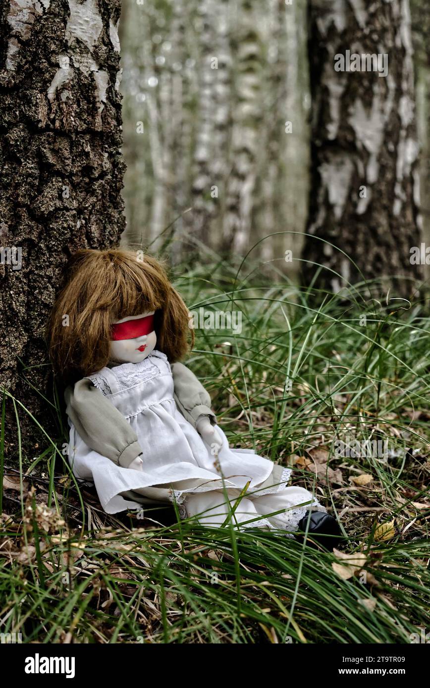Bambola inquietante con ripiegamento cieco a nastro rosso seduta nella foresta. Mistero, thriller, horror, vibrazioni. Foto Stock