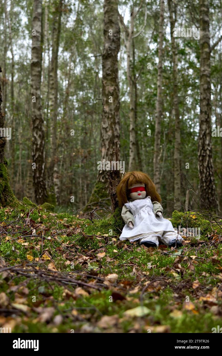Bambola inquietante con ripiegamento cieco a nastro rosso seduta nella foresta. Mistero, thriller, horror, vibrazioni. Foto Stock
