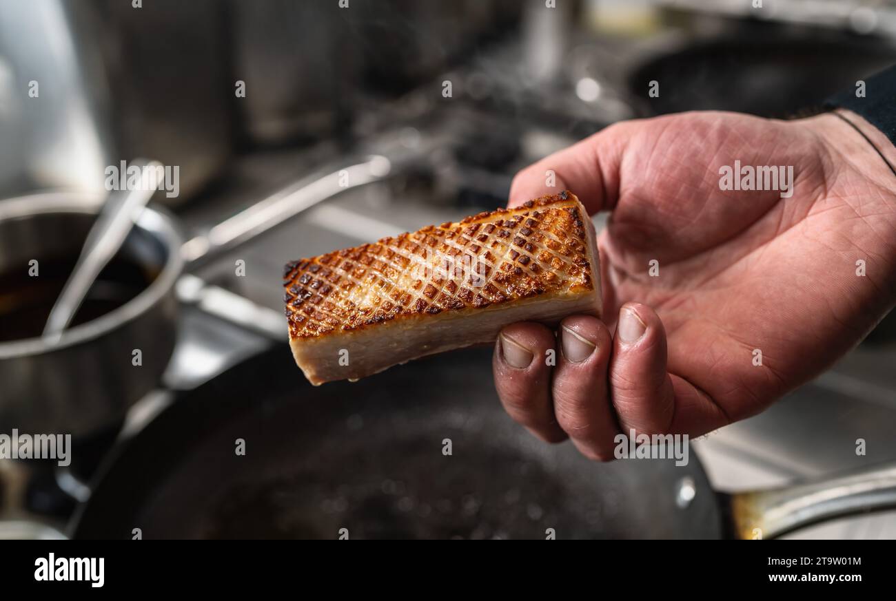 Mano che tiene una pancetta di maiale croccante che è stata arrostita in una padella calda in una cucina professionale in un ristorante. Immagine del concetto di cucina di un hotel di lusso. Foto Stock