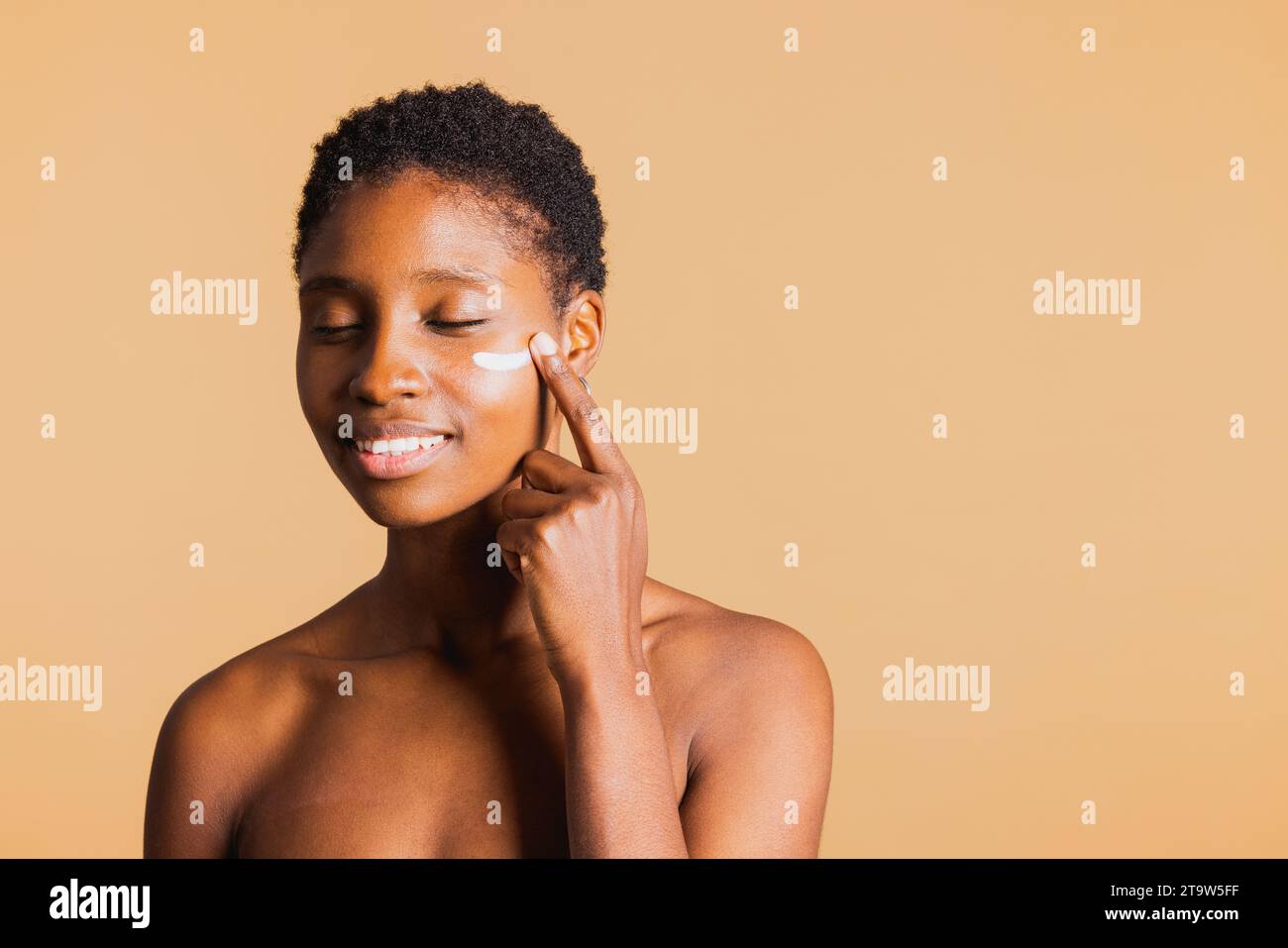 Giovane donna nera sorridente con gli occhi chiusi che applica una crema idratante sul viso. Giovane donna che cura la pelle del viso con un prodotto di bellezza. Foto Stock