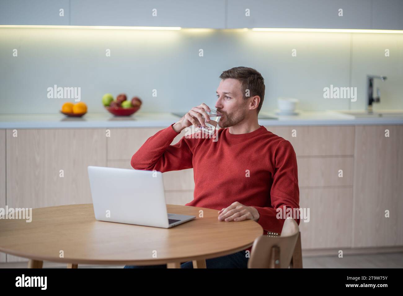 Uomo freelance che beve acqua mentre fa una pausa al lavoro seduto a un tavolo in cucina a casa con un computer portatile. Foto Stock