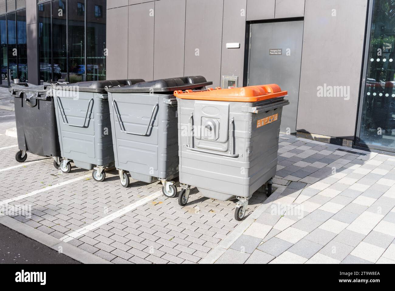 Fila di bidoni usati per riciclare e smaltire rifiuti in un parcheggio sul retro di un moderno edificio per uffici Foto Stock