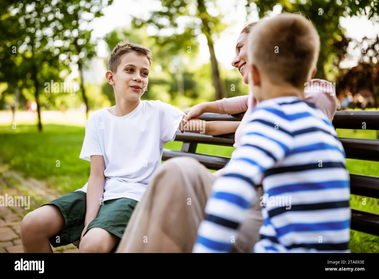 Madre felice è seduta con i suoi figli su panchina nel parco. Stanno parlando e stanno godendo il loro tempo insieme. Foto Stock