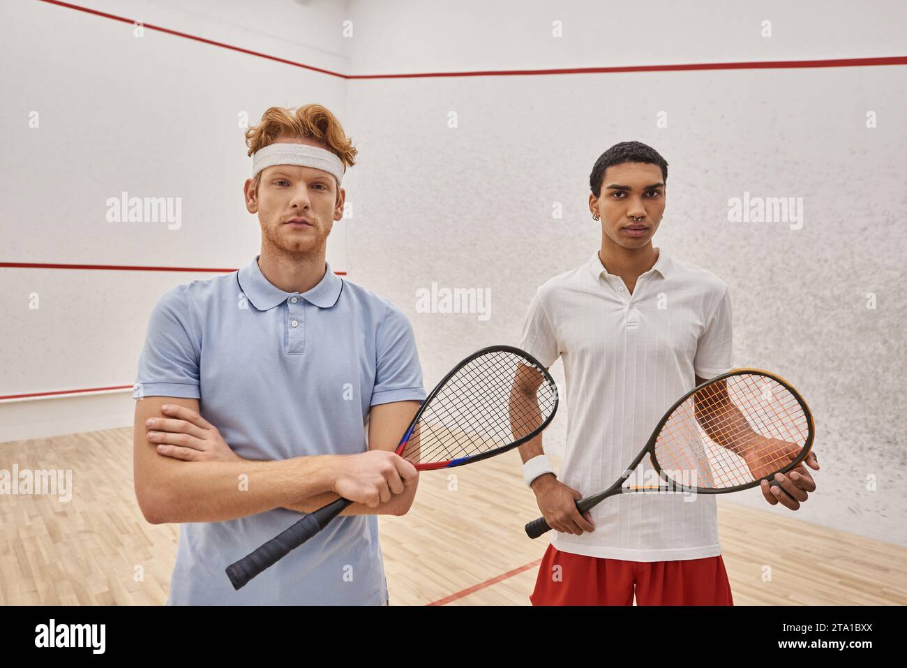 amici maschi interrazziali concentrati in abbigliamento sportivo in piedi insieme alle racchette da squash in campo Foto Stock