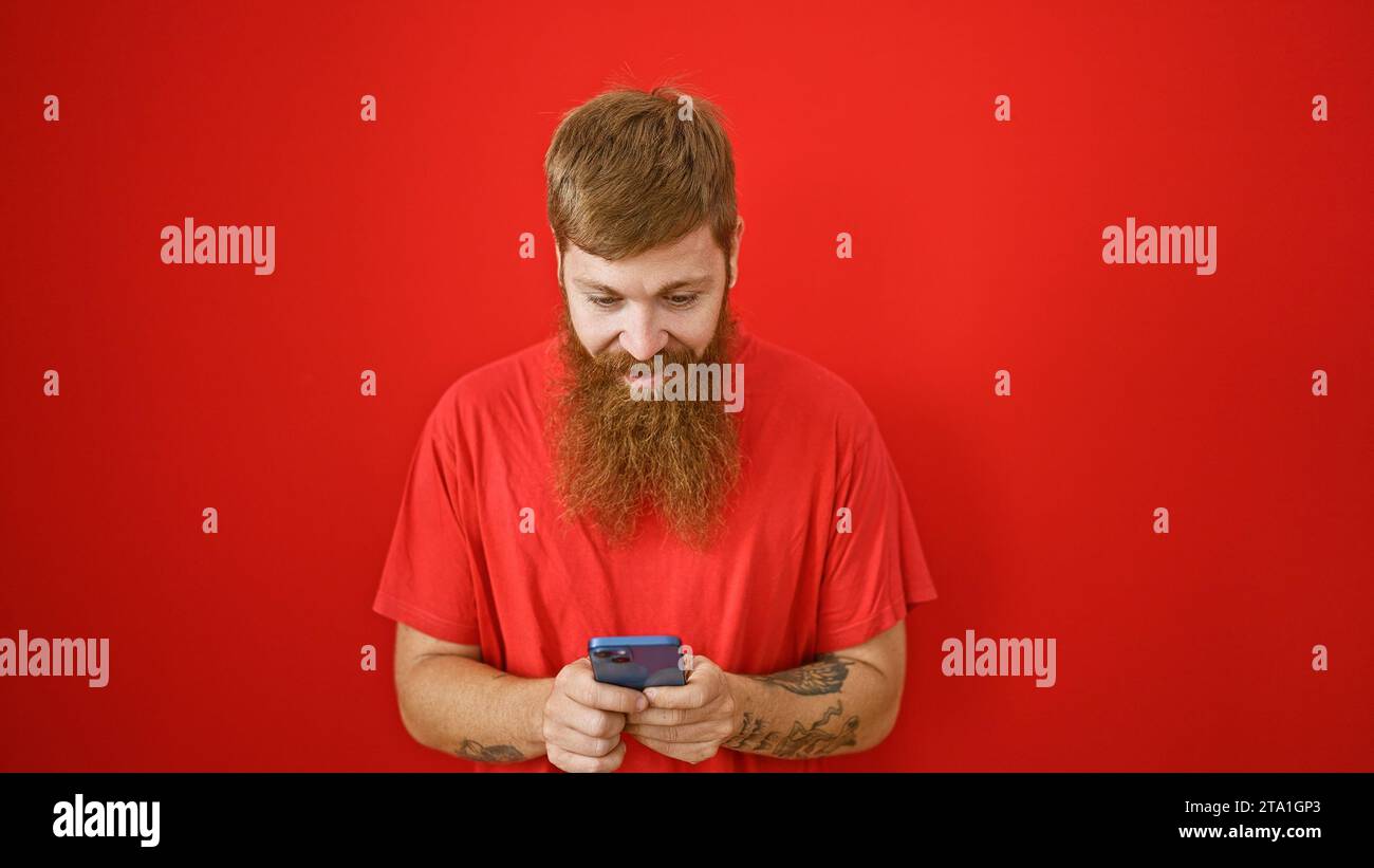 Bellissimo giovane uomo rosso, traboccante di felicità, digitando con sicurezza i messaggi sul suo smartphone su sfondo rosso isolato. la sua risata e.. Foto Stock