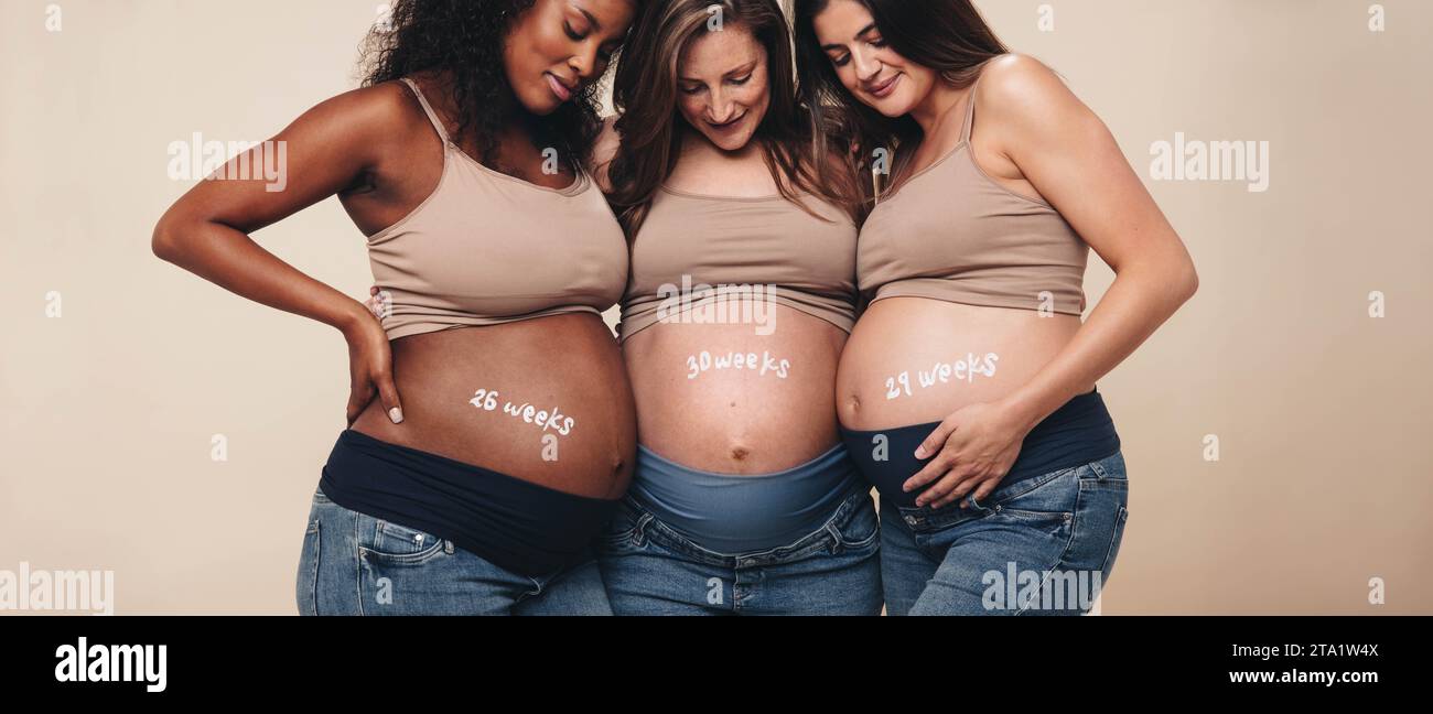 Diverse donne incinte nel terzo trimestre si uniscono, sorridendo ai loro piccoli urti. Si sostengono a vicenda in uno studio, celebrando la madre Foto Stock