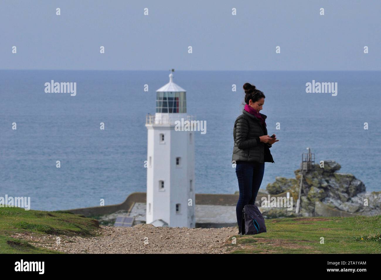 Una giovane donna consulta il suo telefono cellulare, in alto sulle scogliere, con una precedente forma di comunicazione sullo sfondo, la godrevy ligh dal corpo ottagonale Foto Stock