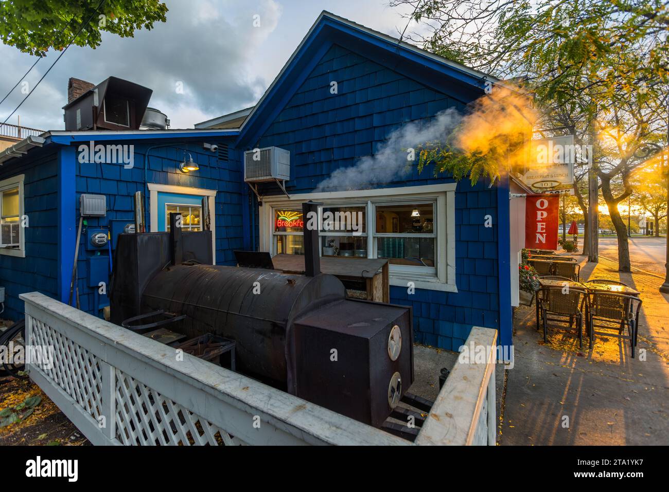 Il fumo sale dal fumoir barbecue fuori dal ristorante Smoke on the Water, con le sue tegole blu, su Park Avenue a Charlevoix, con un'insegna rossa "Aperto" appesa alla porta. Il ristorante per la colazione, noto per i suoi piatti di carne e pesce affumicati, ha aperto nel 2014 in uno degli edifici commerciali più antichi della città. Charlevoix, Michigan, Stati Uniti. Foto Stock