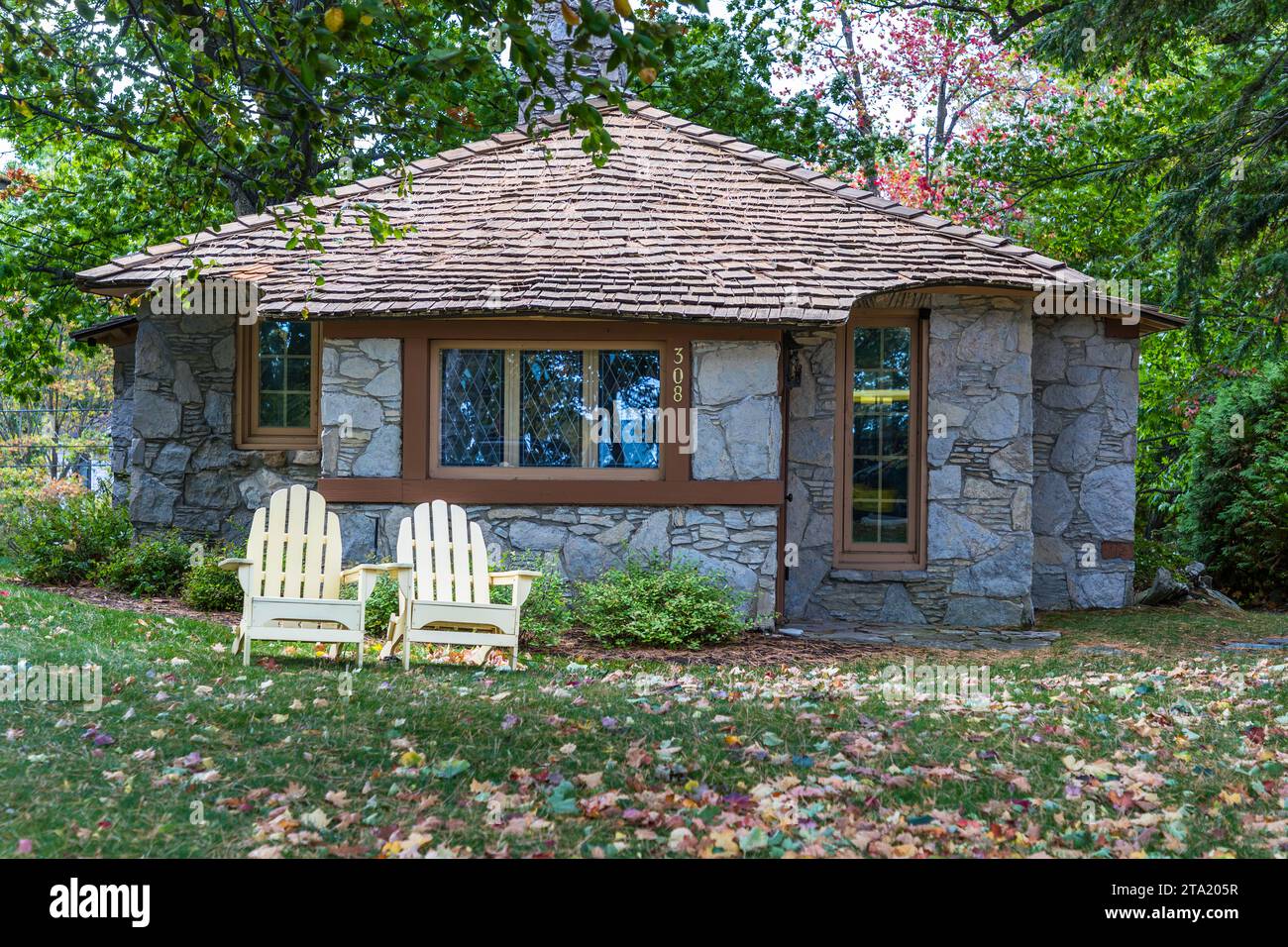 Una piccola casa in pietra con tetto a cono di Earl A. Young con infissi chiari si trova sotto gli alberi a Charlevoix, davanti due sedie Adirondack color crema sul prato. Con i suoi progetti, Young voleva esprimere che una piccola casa in pietra poteva essere impressionante tanto quanto un castello. Charlevoix, Michigan, Stati Uniti. Foto Stock