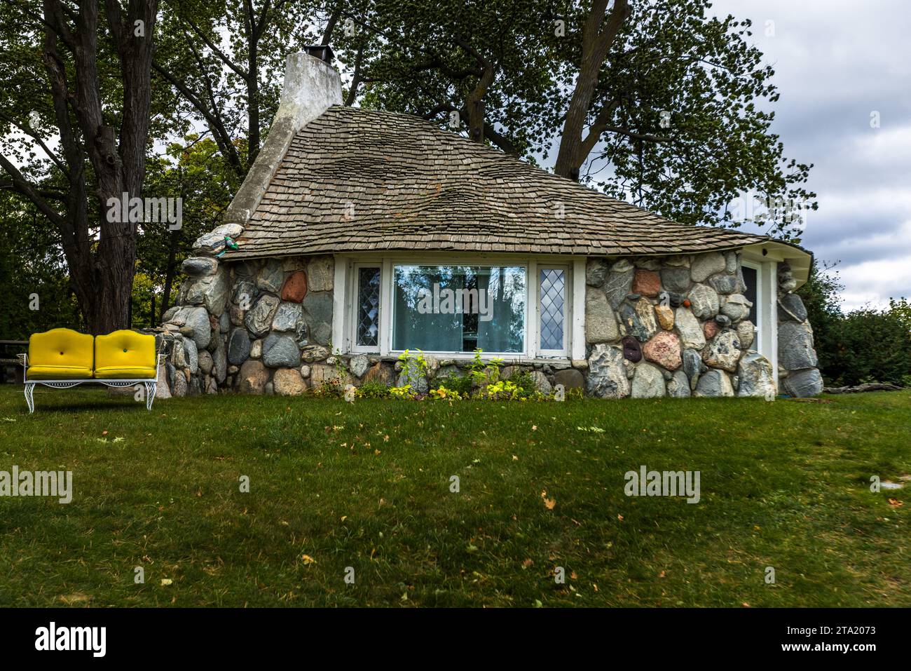 Una Mushroom House di Earl A. Young, il suo tetto curvo in scandole che si inarca sopra una facciata in pietra colorata, si trova nel distretto storico locale di Earl Young a Charlevoix, con una panchina giallo brillante posizionata sul prato davanti. L'architetto ha lavorato principalmente con calcare, pietra di campo e massi che ha trovato nel nord del Michigan. Charlevoix, Michigan, Stati Uniti. Foto Stock
