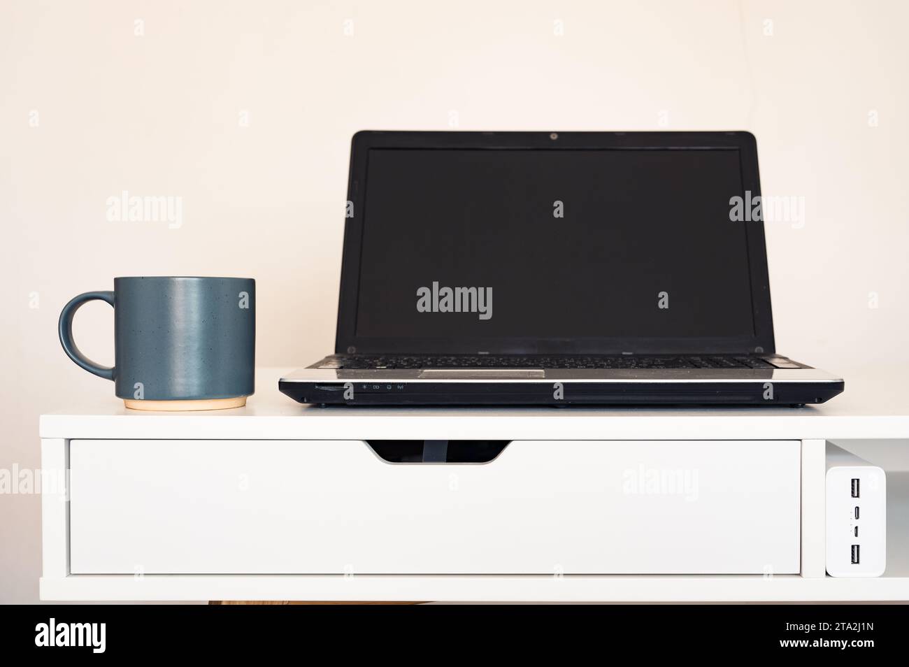 Computer portatile vuoto aperto con mockup e schermo nero su tavolo da scrivania bianco. Tazza con caffè. Caricabatterie. Area di lavoro minimalista Foto Stock