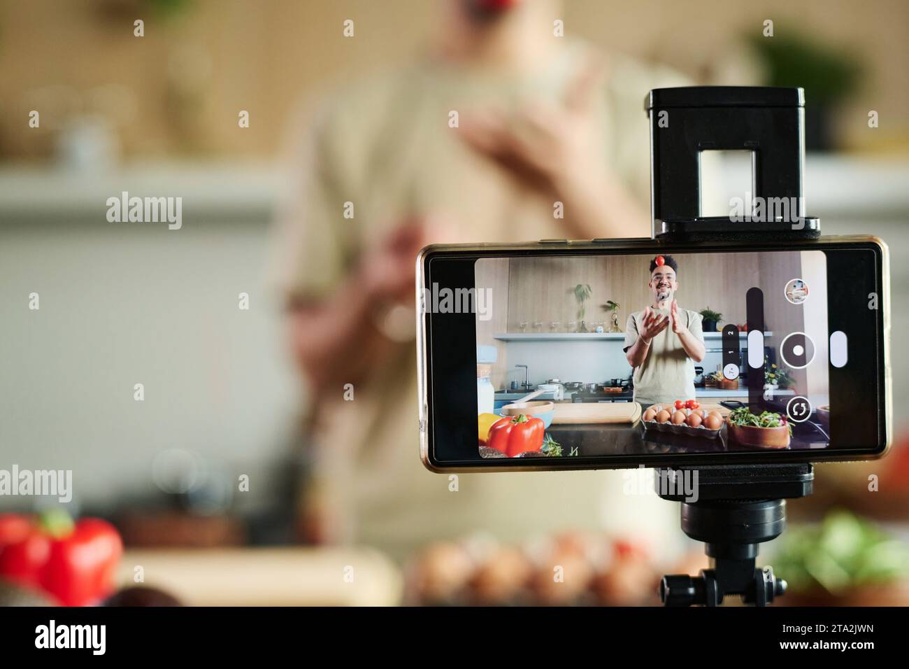 Primo piano sullo smartphone che filma il giovane blogger medio orientale che tosta il pomodoro mentre si trova vicino al tavolo della cucina, si concentra sullo smartphone e sullo spazio di copia Foto Stock