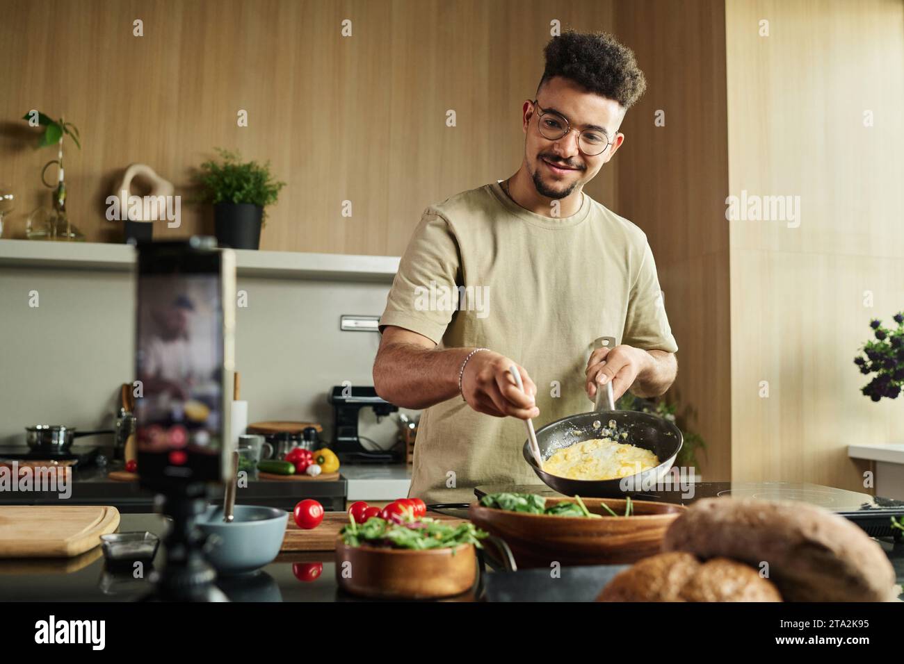 Scatto medio di un giovane influencer medio orientale che tiene in mano PAN con omelette che la mostra alla fotocamera dello smartphone durante la registrazione Foto Stock