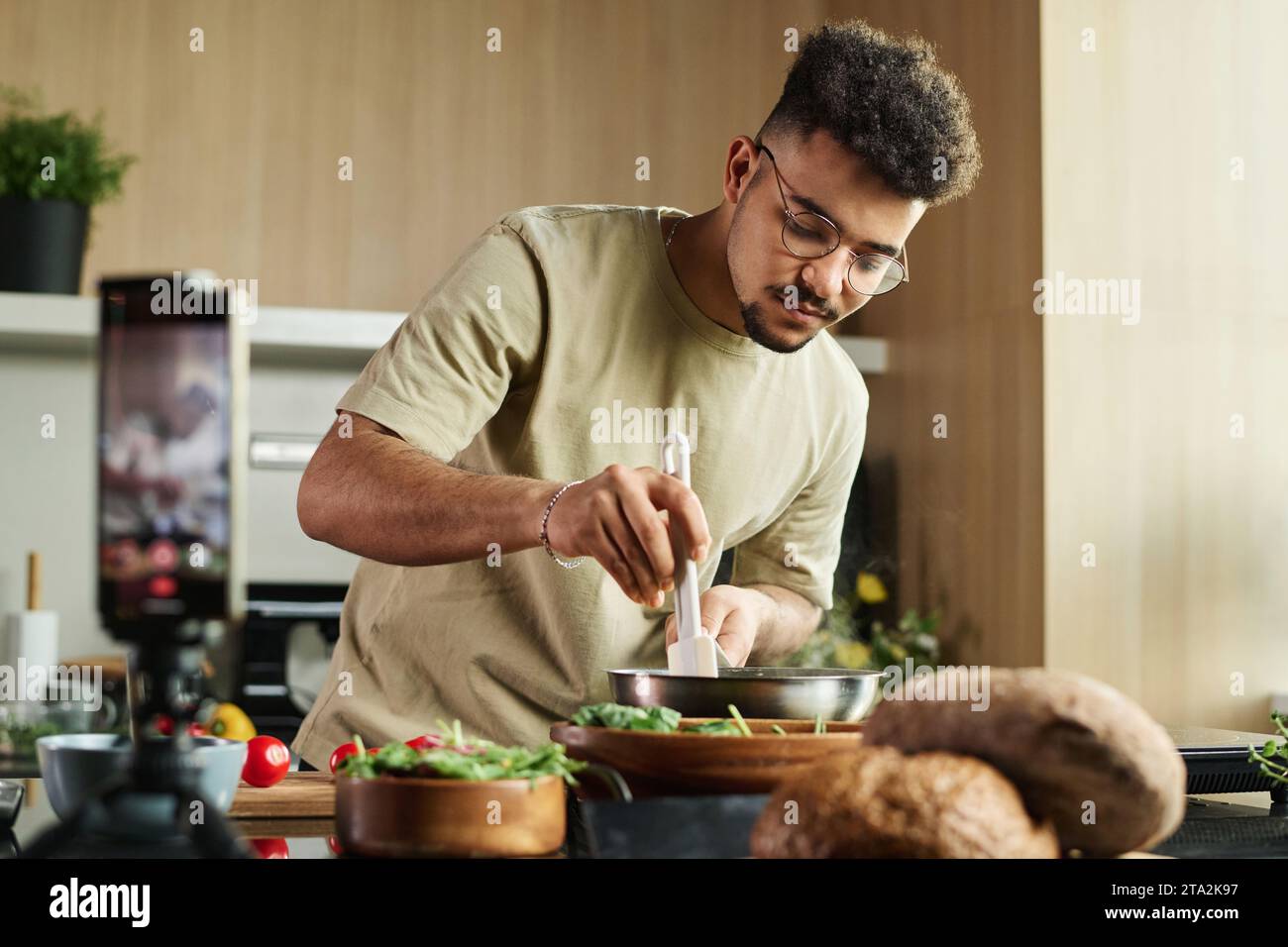 Scatto medio di un giovane chef medio-orientale che mescola il cibo in padella con la spatola durante la realizzazione di un video sullo smartphone Foto Stock