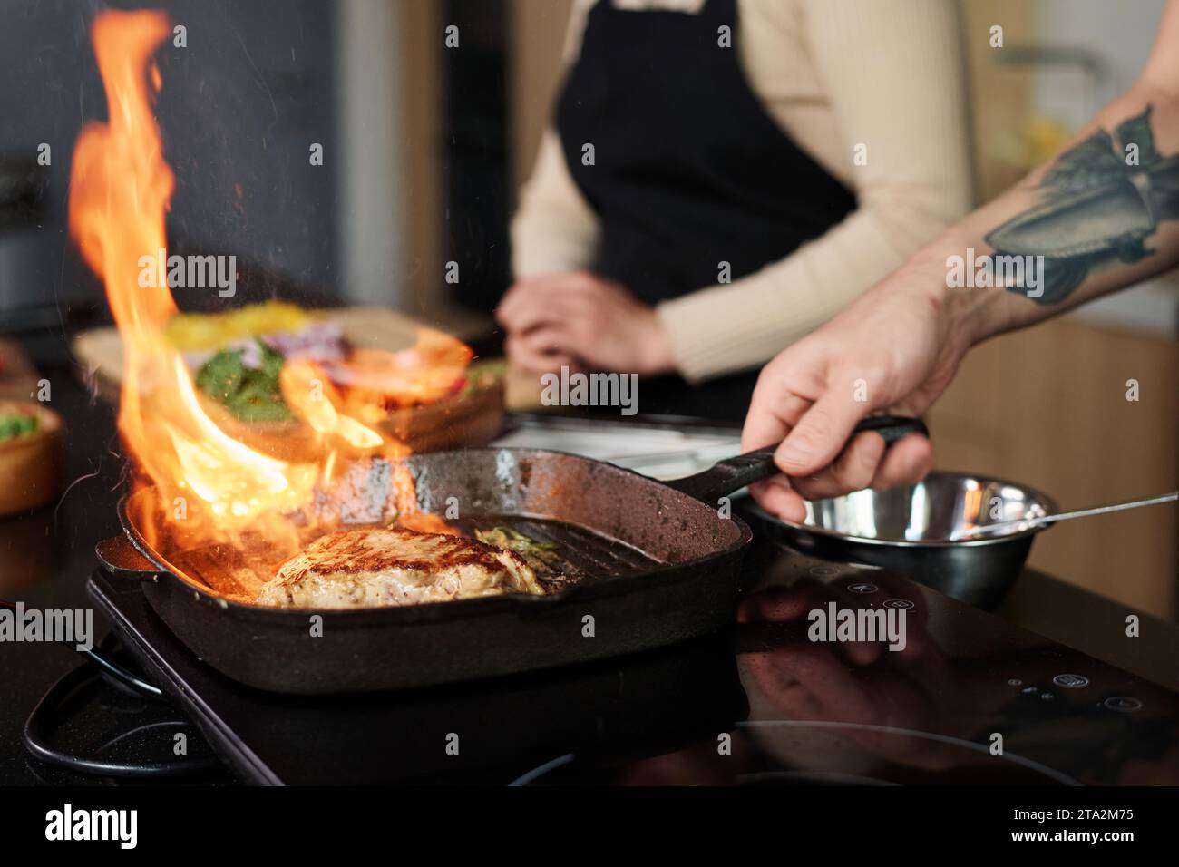 Primo piano di un uomo irriconoscibile che frigge bistecca in padella alla griglia ad alte temperature con una donna sconosciuta sullo sfondo, concentrati sulla carne Foto Stock