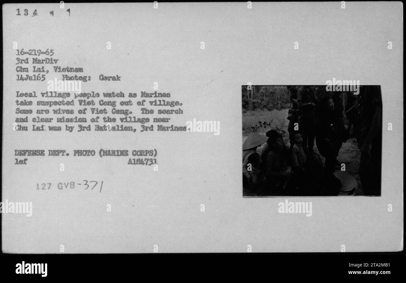 Gli abitanti del villaggio osservano mentre i marines catturano sospetti insorti Viet Cong nel loro villaggio vicino a Chu Lai, in Vietnam. Questa operazione ebbe luogo il 14 luglio 1965, condotta dal 3rd Battalion, 3rd Marines della 3rd Marine Division. La fotografia cattura la ricerca e la missione chiara, con alcuni degli osservatori che sono le mogli dei membri dei Viet Cong. Foto Stock