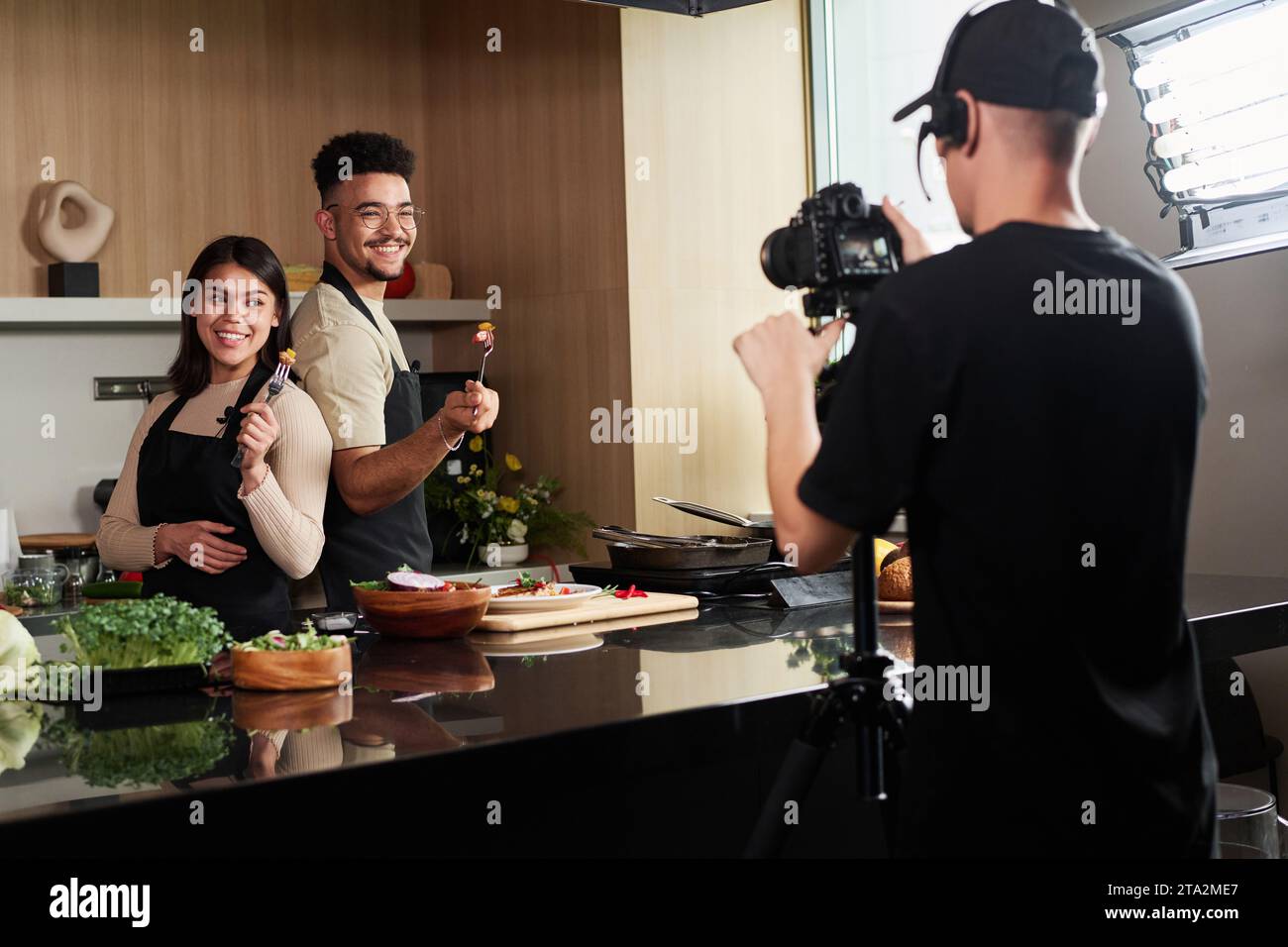 Uomo medio-orientale e donna ispanica in piedi dietro tenendo forchette con il cibo dal bancone della cucina mentre viene fotografato dal cameraman Foto Stock