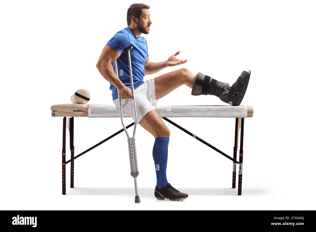 Giocatore di football con una stampella e un piede ferito seduto su un letto di terapia fisica isolato su sfondo bianco Foto Stock
