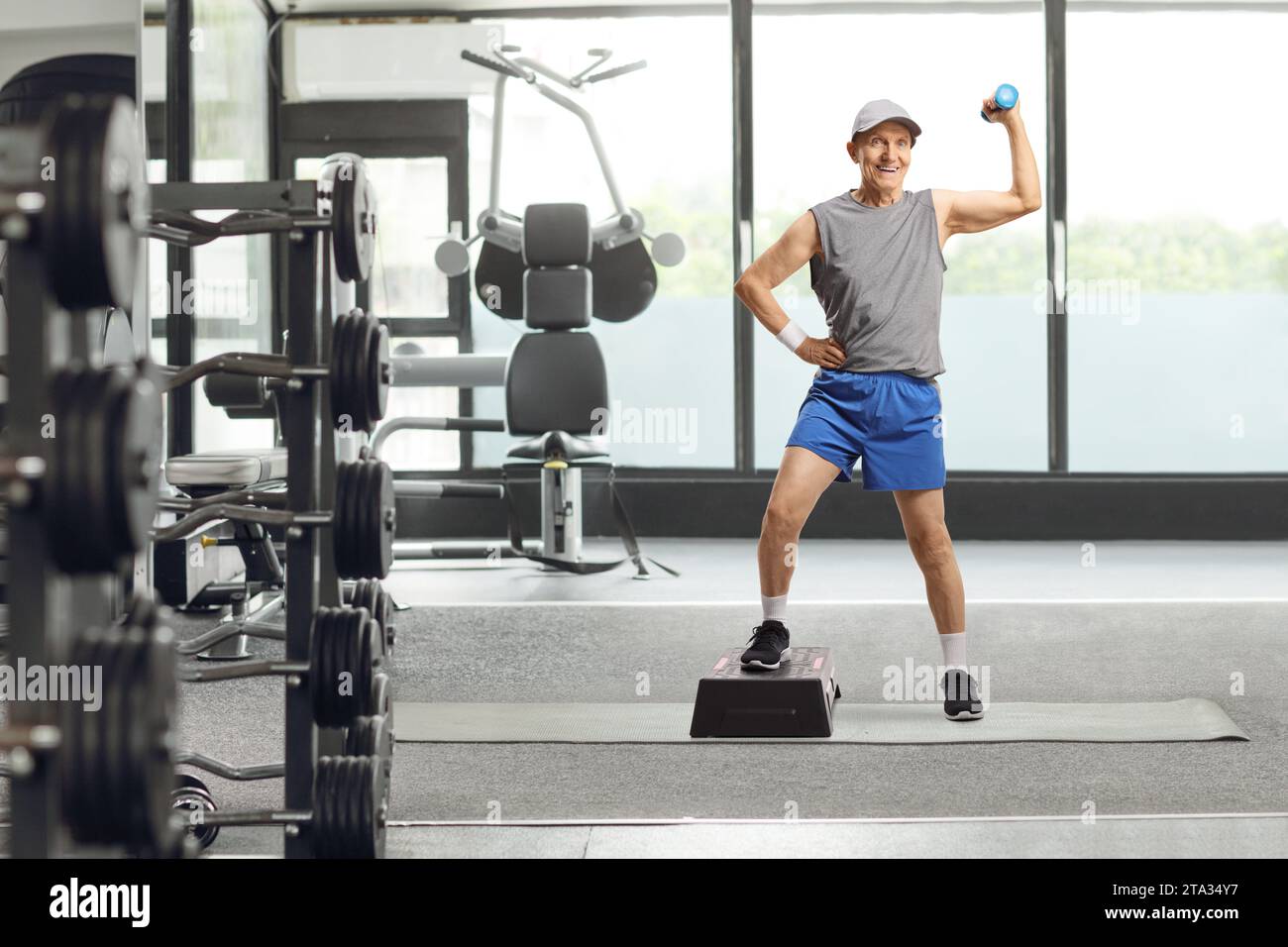 Ritratto a figura intera di un uomo anziano in abbigliamento sportivo che si esercita con una pedana aerobica a gradini e un piccolo peso in palestra Foto Stock