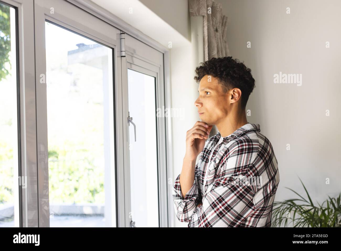 Ritratto di un uomo birazziale concentrato che guarda attraverso la finestra, tenendo il mento con la mano a casa, copiando lo spazio. Espressione, tempo libero, gesticolazione e l Foto Stock
