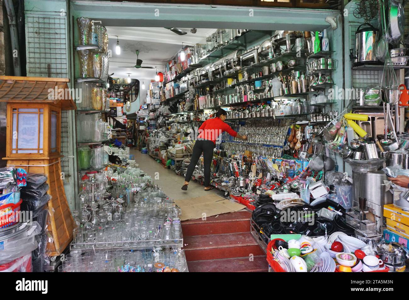 Un commerciante lavora nel suo negozio, rifornito dal pavimento al soffitto e traboccante sul marciapiede con piatti, bicchieri e molto altro ad Hanoi, in Vietnam Foto Stock