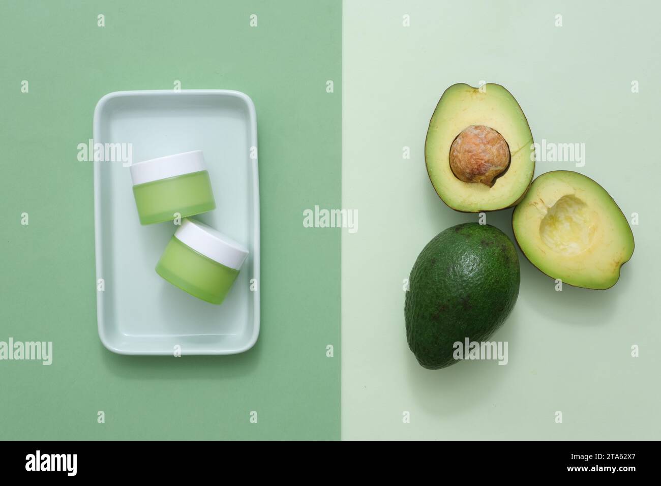 Scena minimalista con ingrediente di avocado e barattolo cosmetico senza marchio per la pubblicità. Vista dall'alto di metà di avocado su sfondo verde chiaro e crema Foto Stock