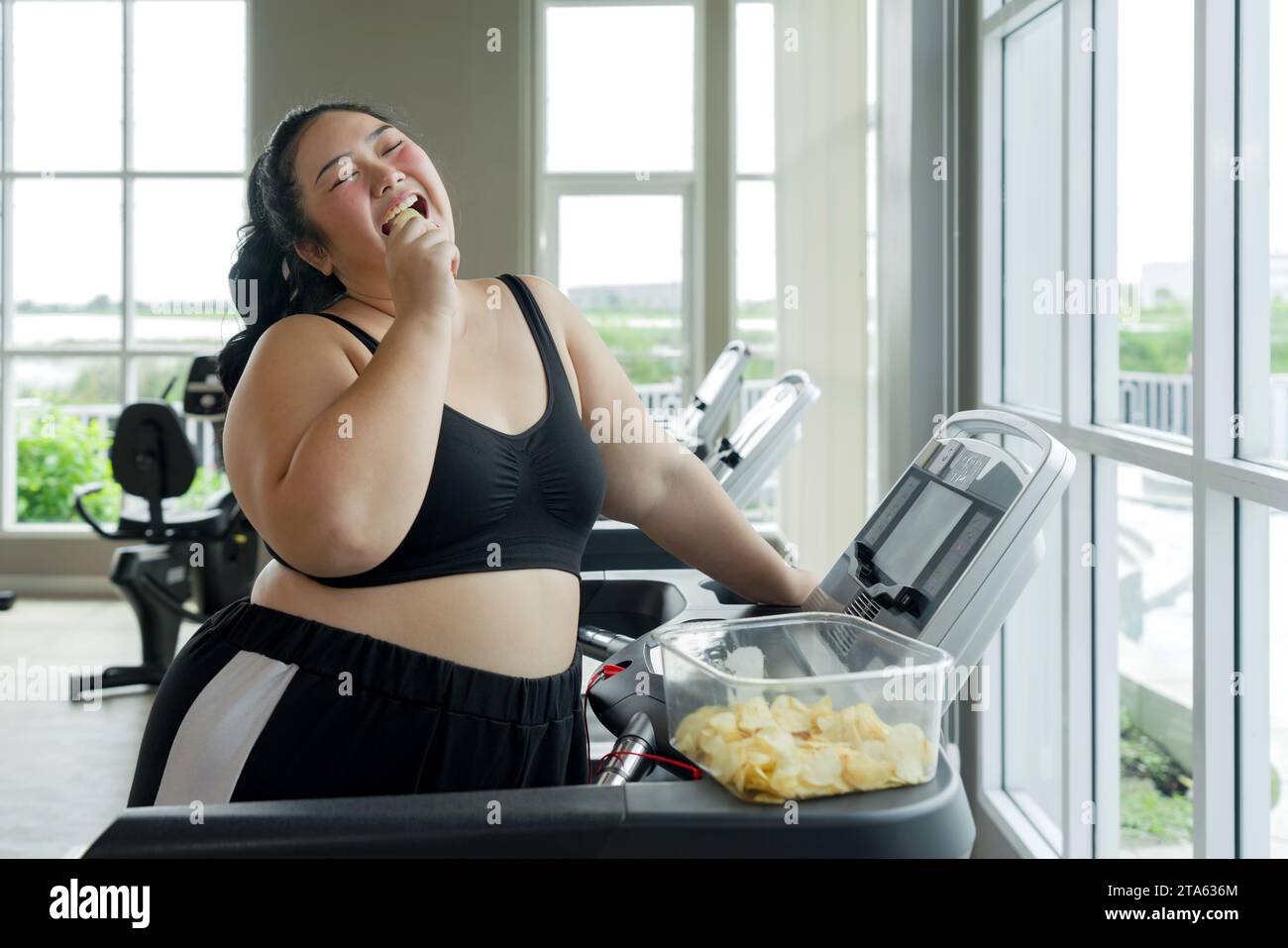 Una donna maggiorenne sta mangiando patatine mentre si allena su un tapis roulant in palestra, combinando il piacere con il fitness. Foto Stock