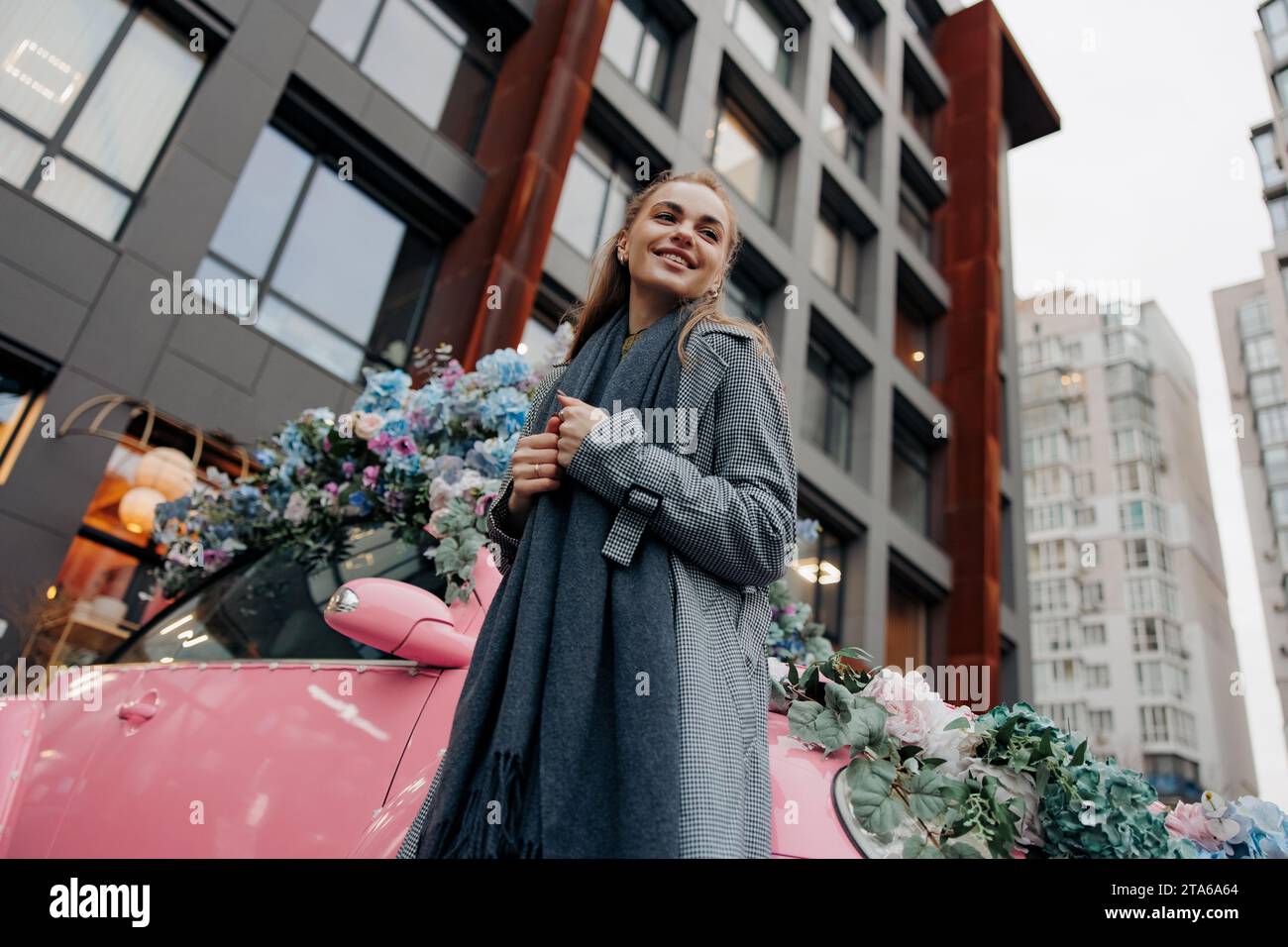 Giovane donna sorridente felice in piedi vicino a un'auto rosa decorata da ghirlande di fiori sulla strada della città sullo sfondo di edifici. Vista dal basso. Foto Stock