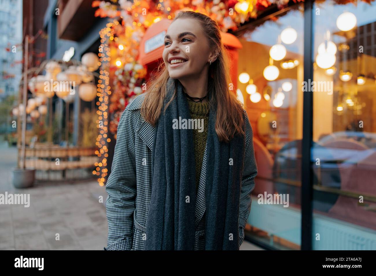 Giovane donna sorridente e felice che cammina per la strada della città decorata da ghirlande luminose di vacanza sullo sfondo di edifici. Vista ravvicinata. Foto Stock