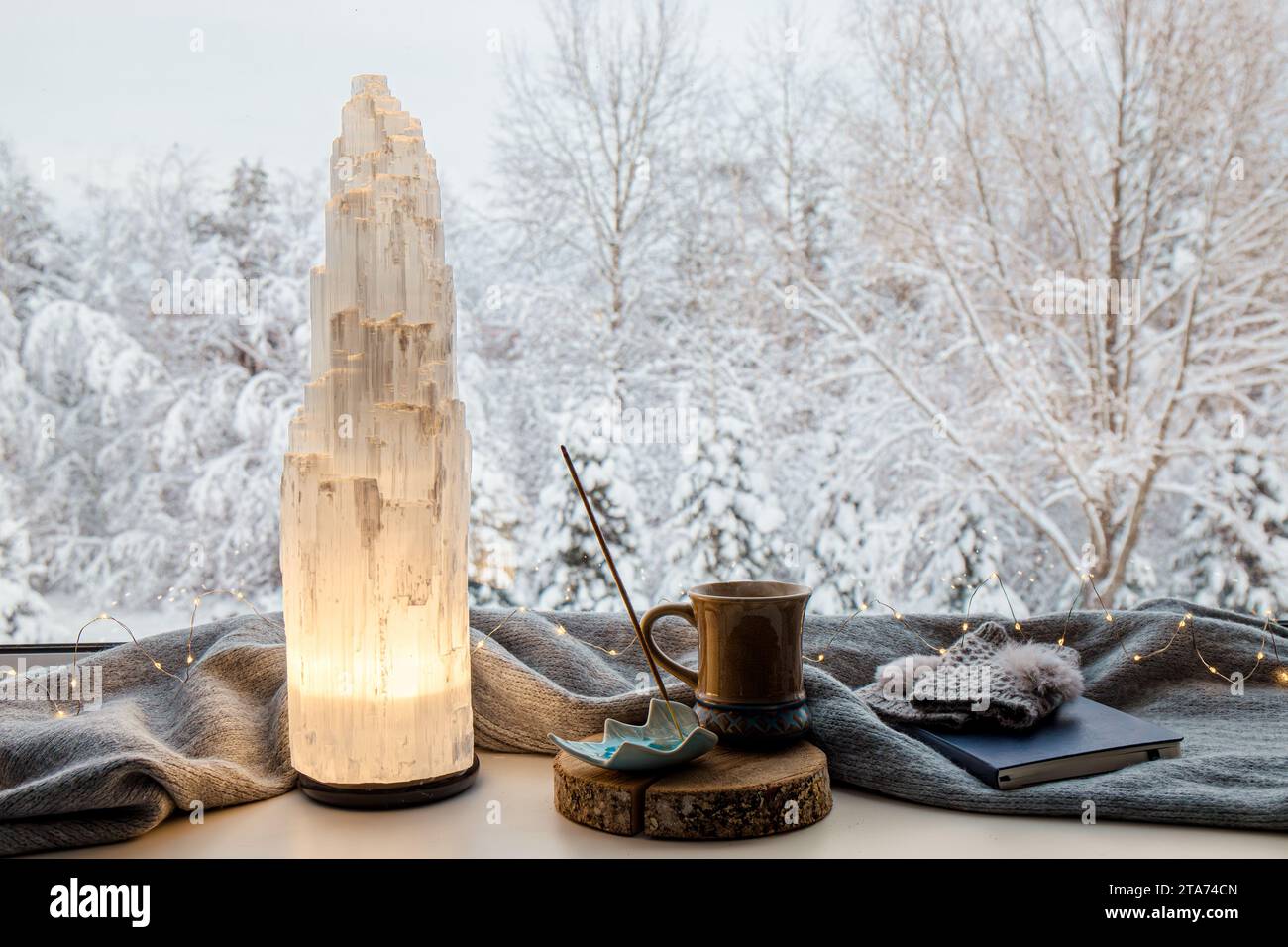 Accogliente casa invernale con lampada elettrica a torre in cristallo selenite naturale illuminata su davanzale, sciarpa e guanti di colore grigio, quaderno di carta, tè. Foto Stock