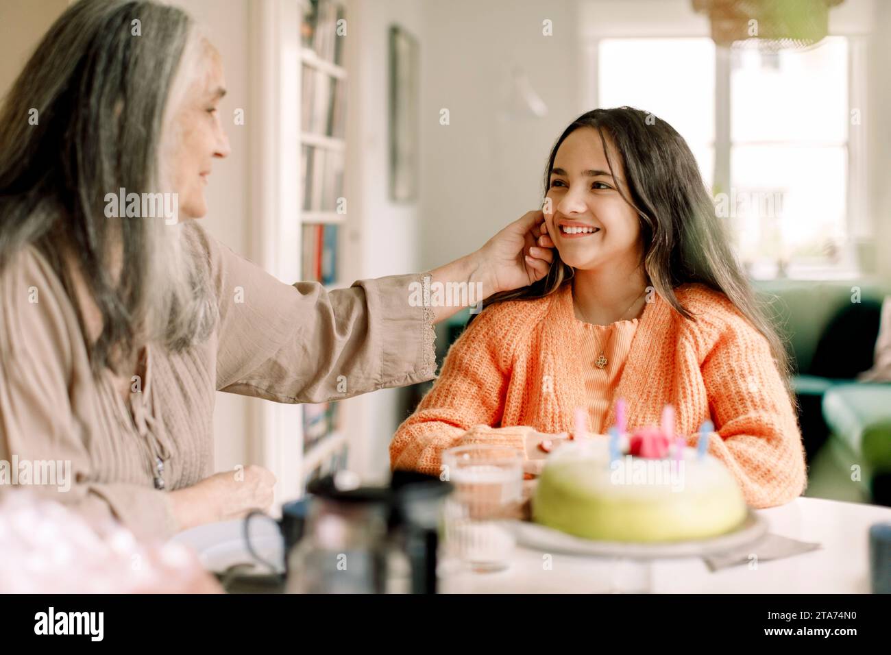 Donna anziana che tocca la guancia della nipote seduta con una torta di compleanno al tavolo da pranzo Foto Stock