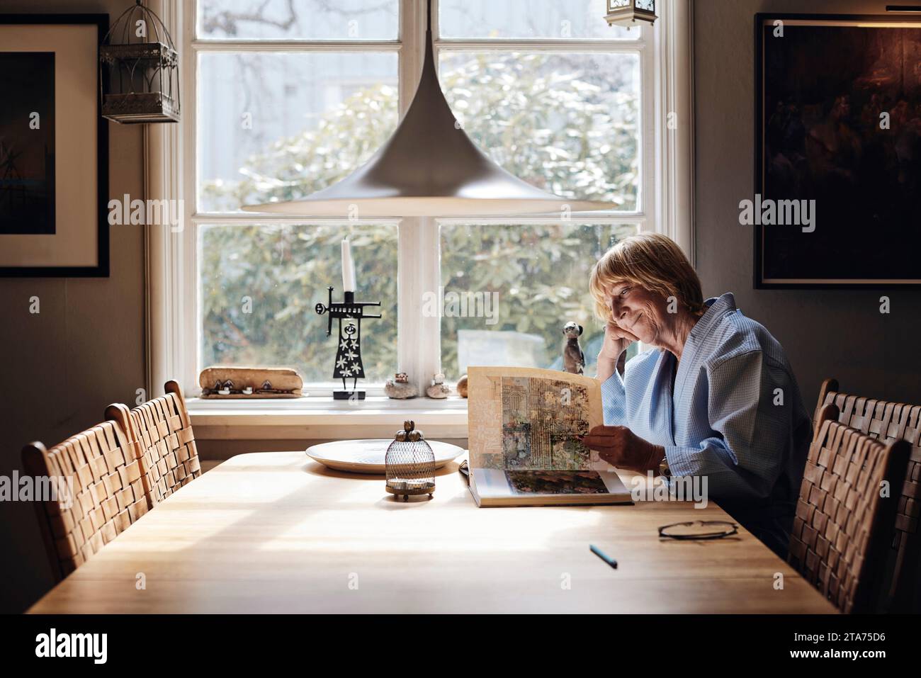Donna anziana sorridente che guarda un libro fotografico sul tavolo da pranzo a casa Foto Stock