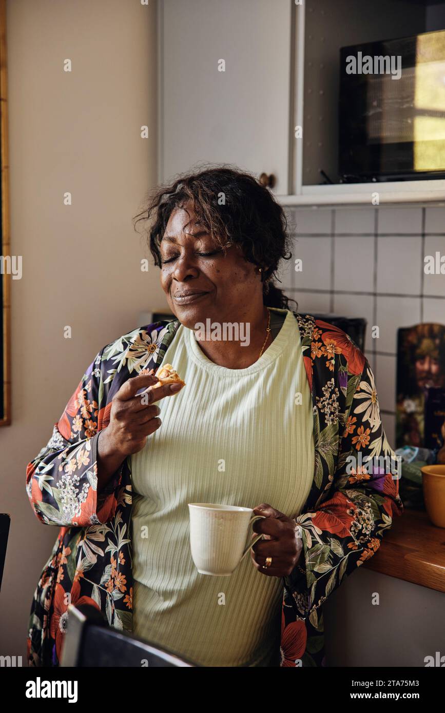 Donna matura con gli occhi chiusi che beve biscotti e caffè in cucina a casa Foto Stock