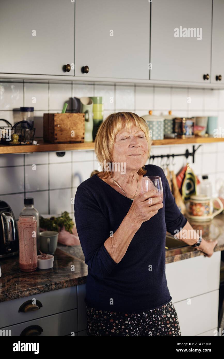 Donna anziana bionda con gli occhi chiusi che beve un bicchiere in cucina a casa Foto Stock