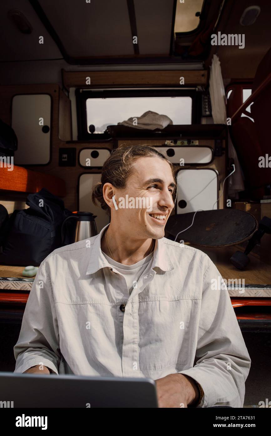 Uomo sorridente che guarda lontano, indossa cuffie auricolari wireless mentre è seduto vicino al furgone Foto Stock