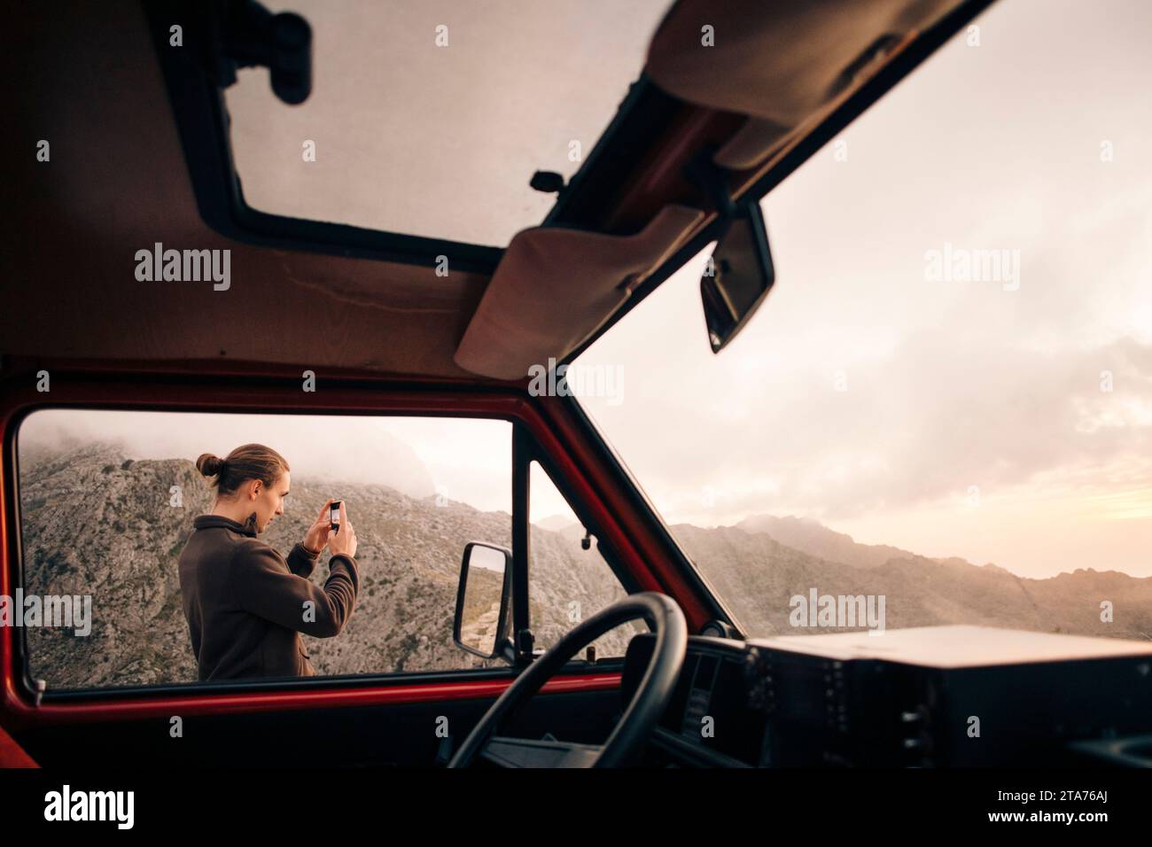 Giovane che fotografa le montagne attraverso uno smartphone visto dalla finestra del furgone in vacanza Foto Stock