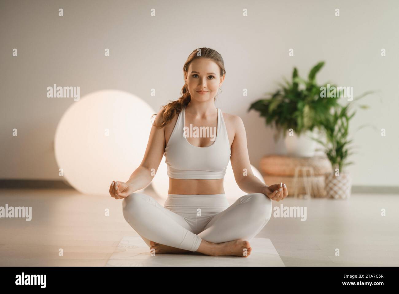 Ritratto di una ragazza in abiti bianchi seduta in posizione di loto su un tappetino durante una lezione di yoga al coperto. Foto Stock