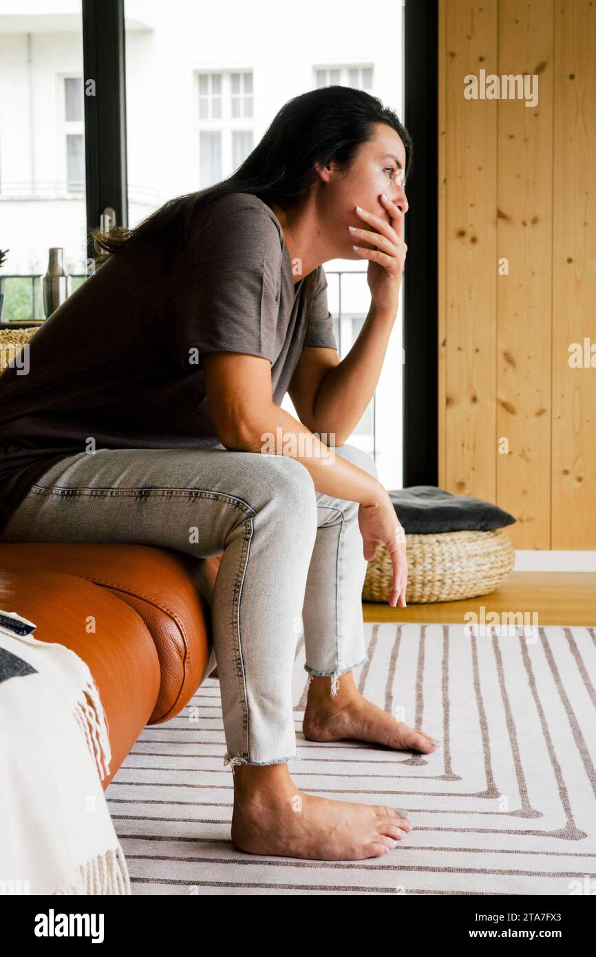 Donna preoccupata con la bocca che copre le mani seduta sul divano a casa Foto Stock