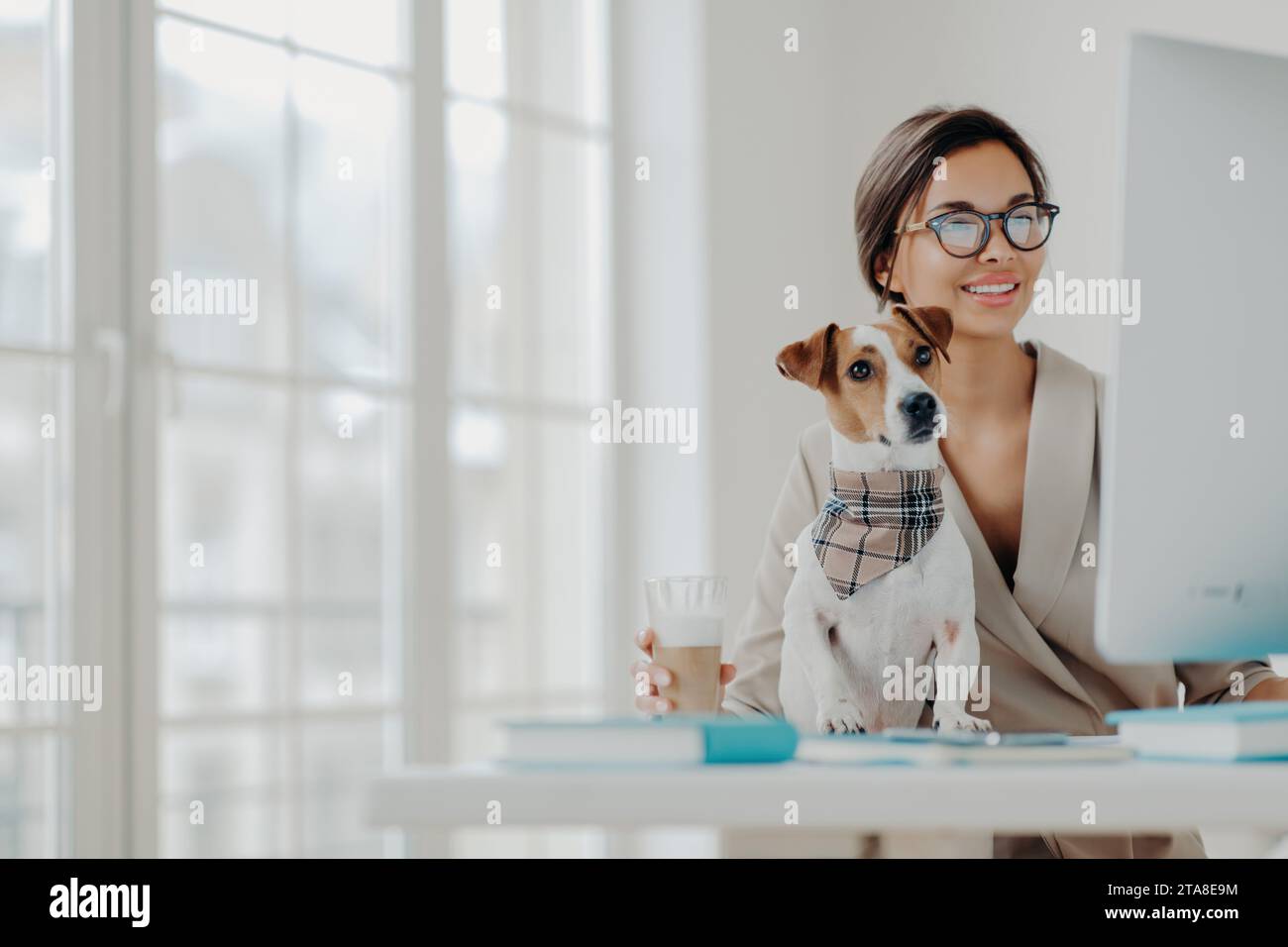 Donna professionale che lavora al computer con il suo cane, elegante spazio ufficio, che esemplifica l'equilibrio tra lavoro e vita privata Foto Stock