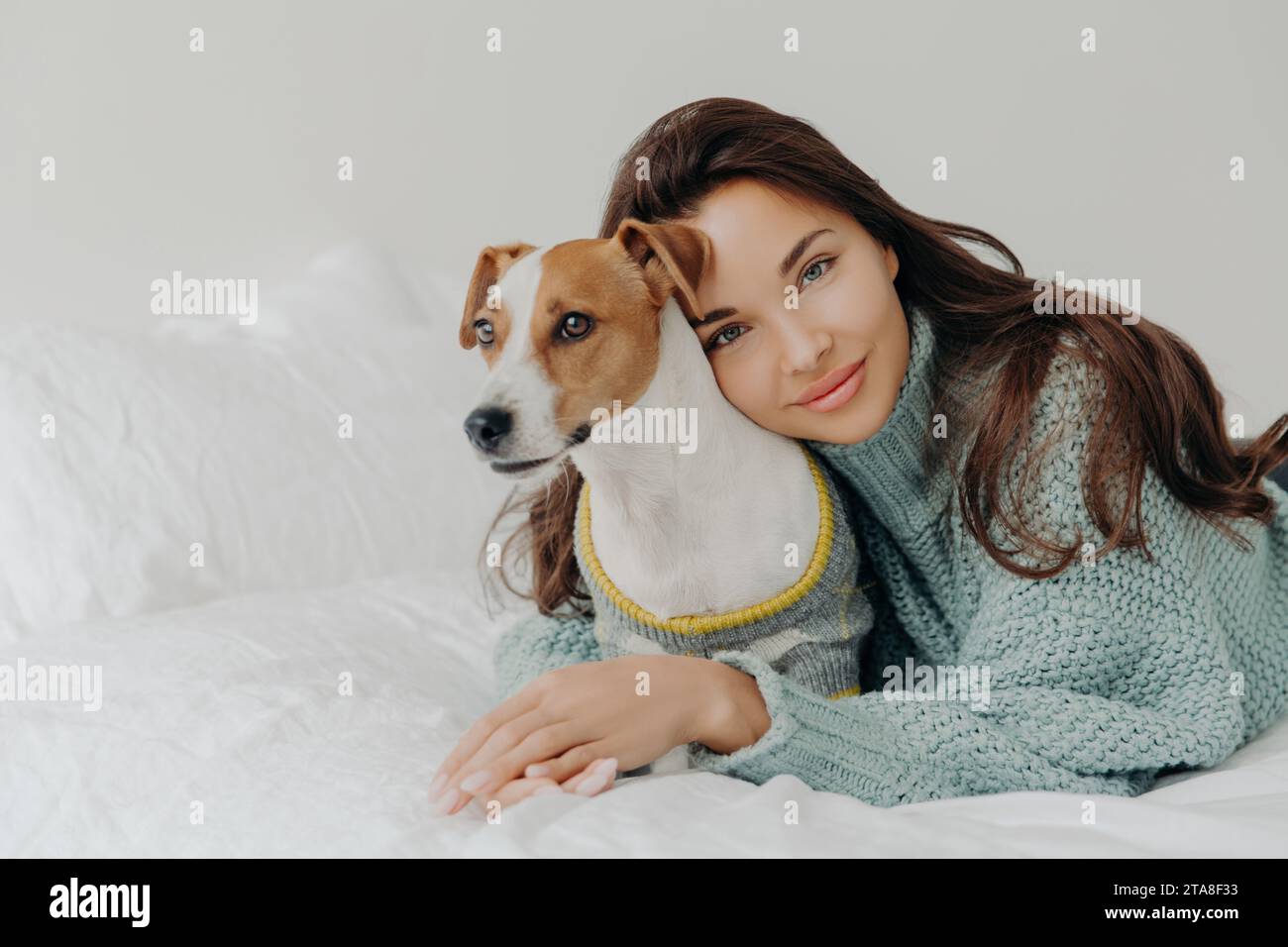 Donna rilassata con un sorriso sereno, sdraiata accanto al suo cane su un letto bianco, evocando un senso di pace e compagnia Foto Stock