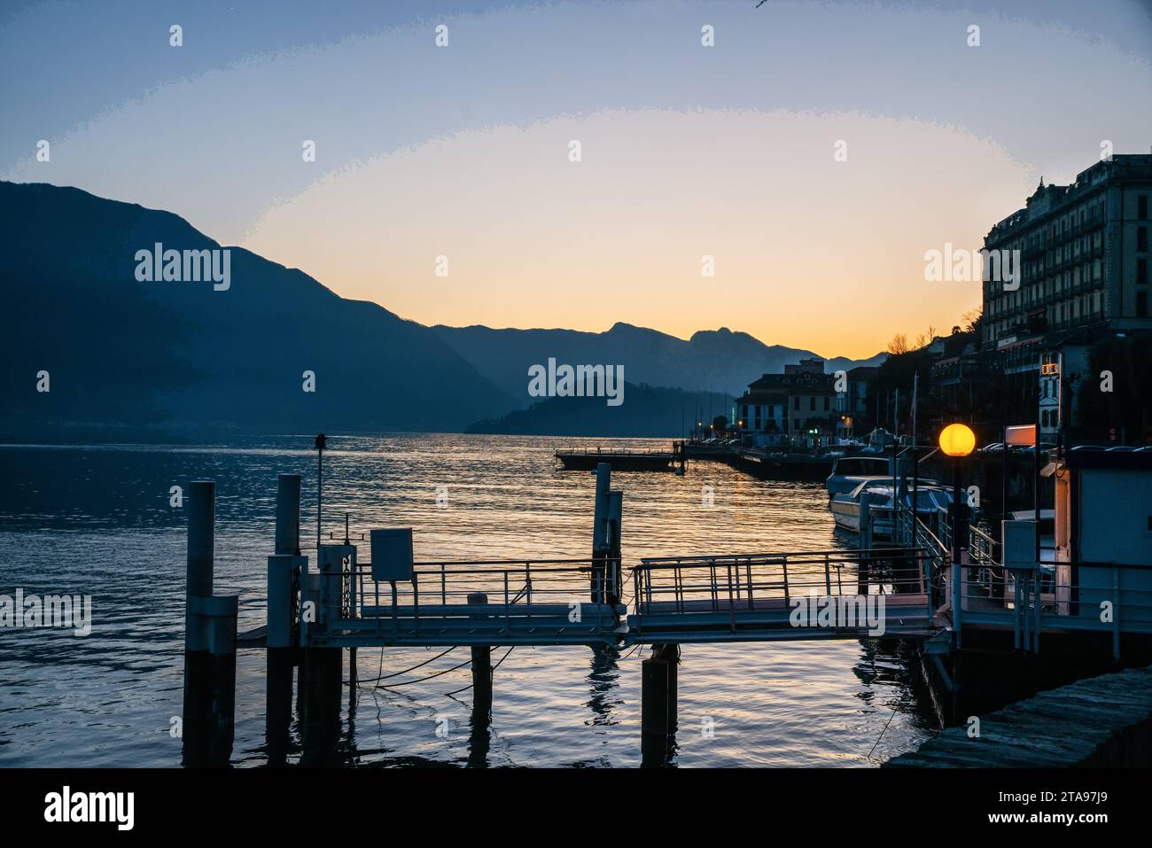 La vista serale del tramonto sul lago di Como e alcune barche attraccano e gli edifici della costa (hotel?). Il molo e la luce elettrica. Breve viaggio a Natale Foto Stock