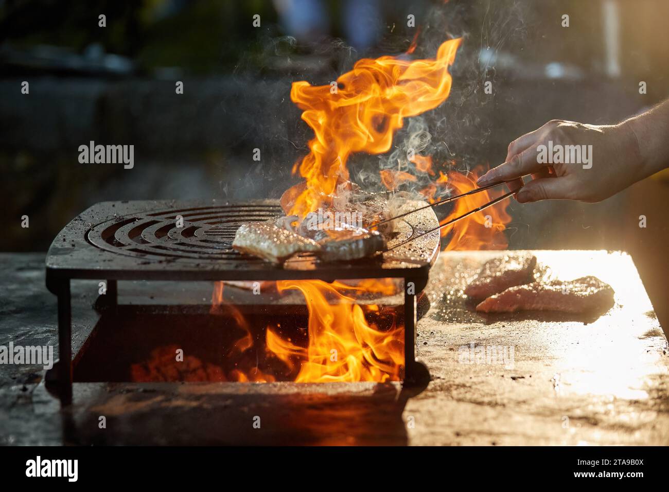 Uno chef prepara una bistecca su un grill all'aperto. Primo piano della cucina al festival del giardino, fiamme, profondità di campo bassa, sfondo sfocato molto colorato, Foto Stock