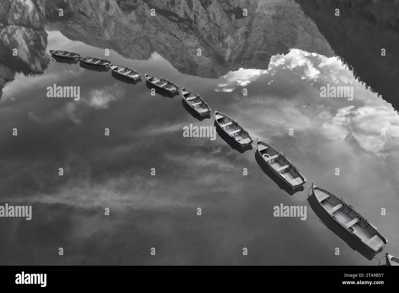 Vista monocromatica, artistica e perpendicolare di barche in legno in fila tra cime e cielo speculare sul lago di Braies nelle Dolomiti. Foto Stock