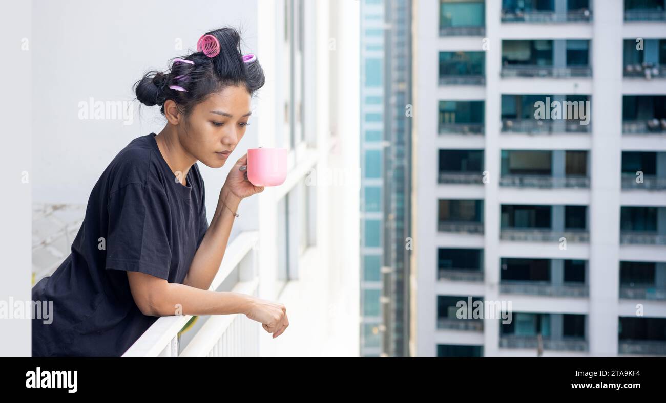 Una donna in curler tiene una tazza sul balcone di un grattacielo in una zona residenziale Foto Stock