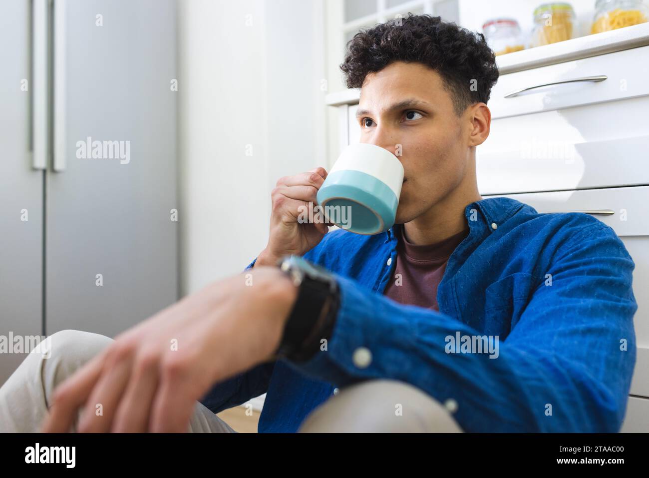 Un uomo birazziale seduto sul pavimento e bevuto il tè in cucina a casa, lo spazio per le fotocopie. Espressione, cibo e bevande e vita domestica inalterata. Foto Stock