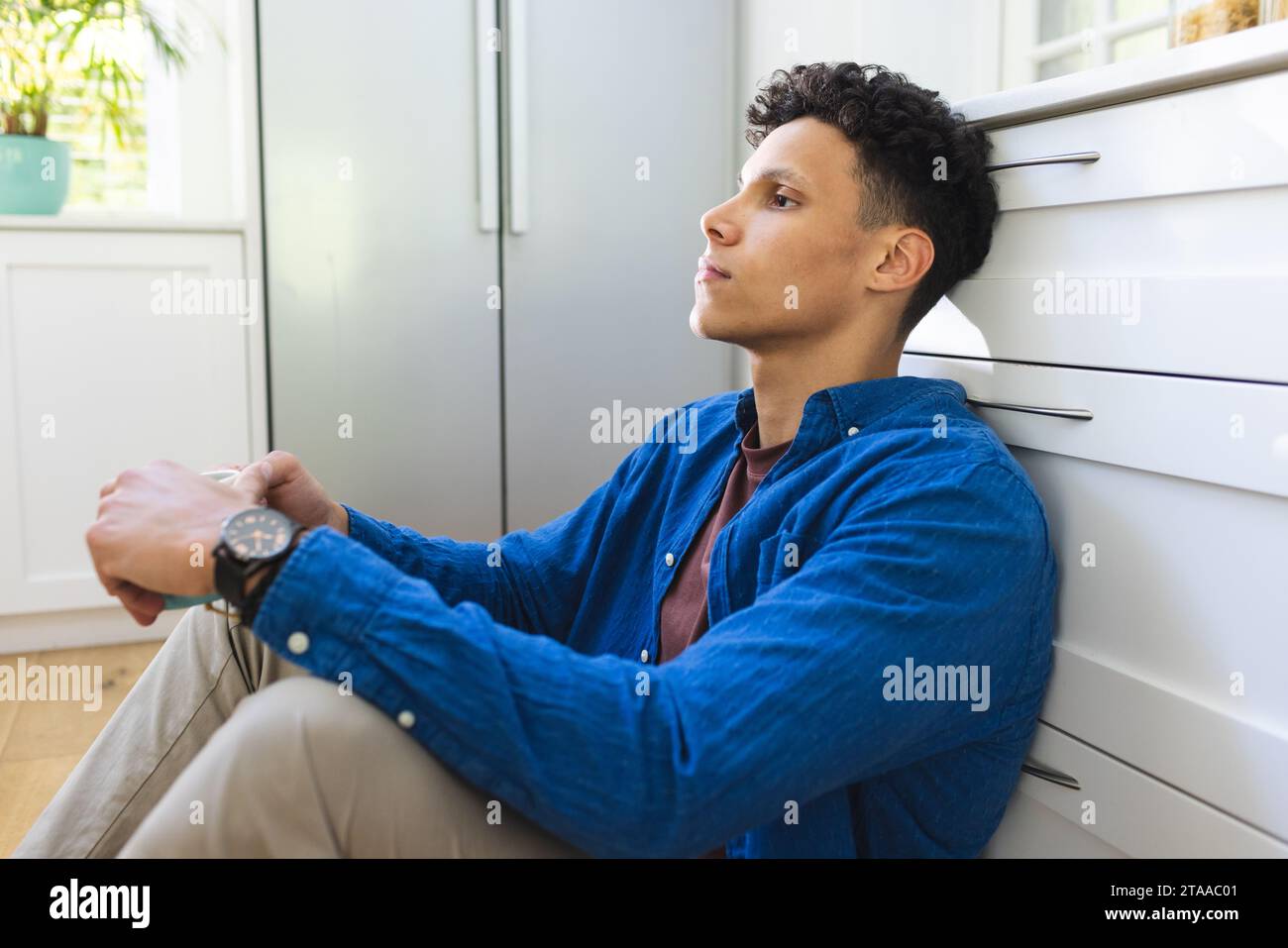 Un uomo birazziale seduto sul pavimento e tenuto una tazza di tè in cucina a casa, lo spazio per le copie. Espressione, cibo e bevande e vita domestica inalterata. Foto Stock