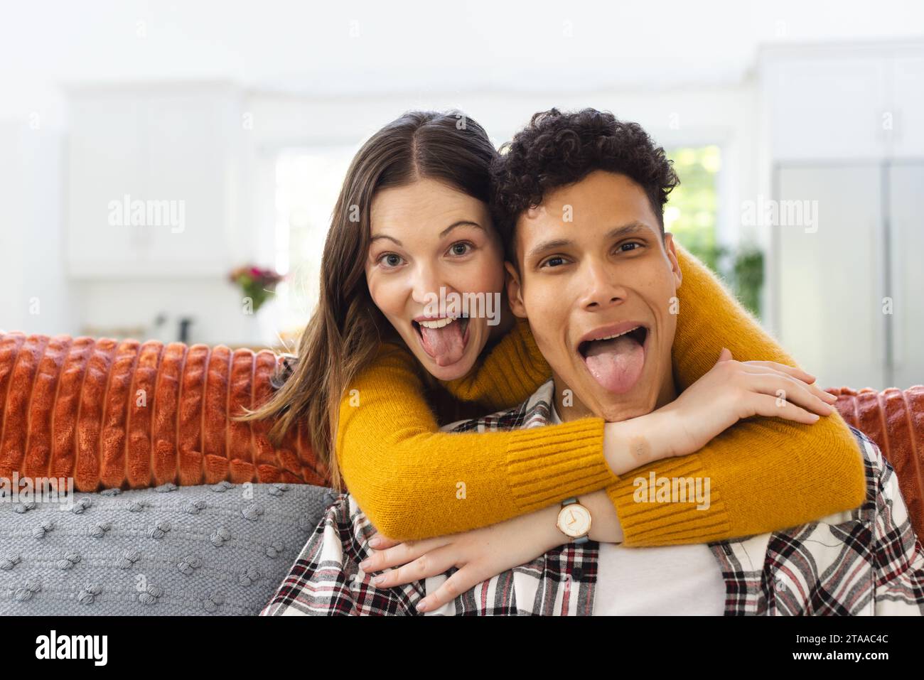 Ritratto di una coppia felice e variegata che si abbraccia sul divano e fa facce divertenti a casa, spazio copia. Espressione, tempo libero, amore, insieme, gesticolazione Foto Stock