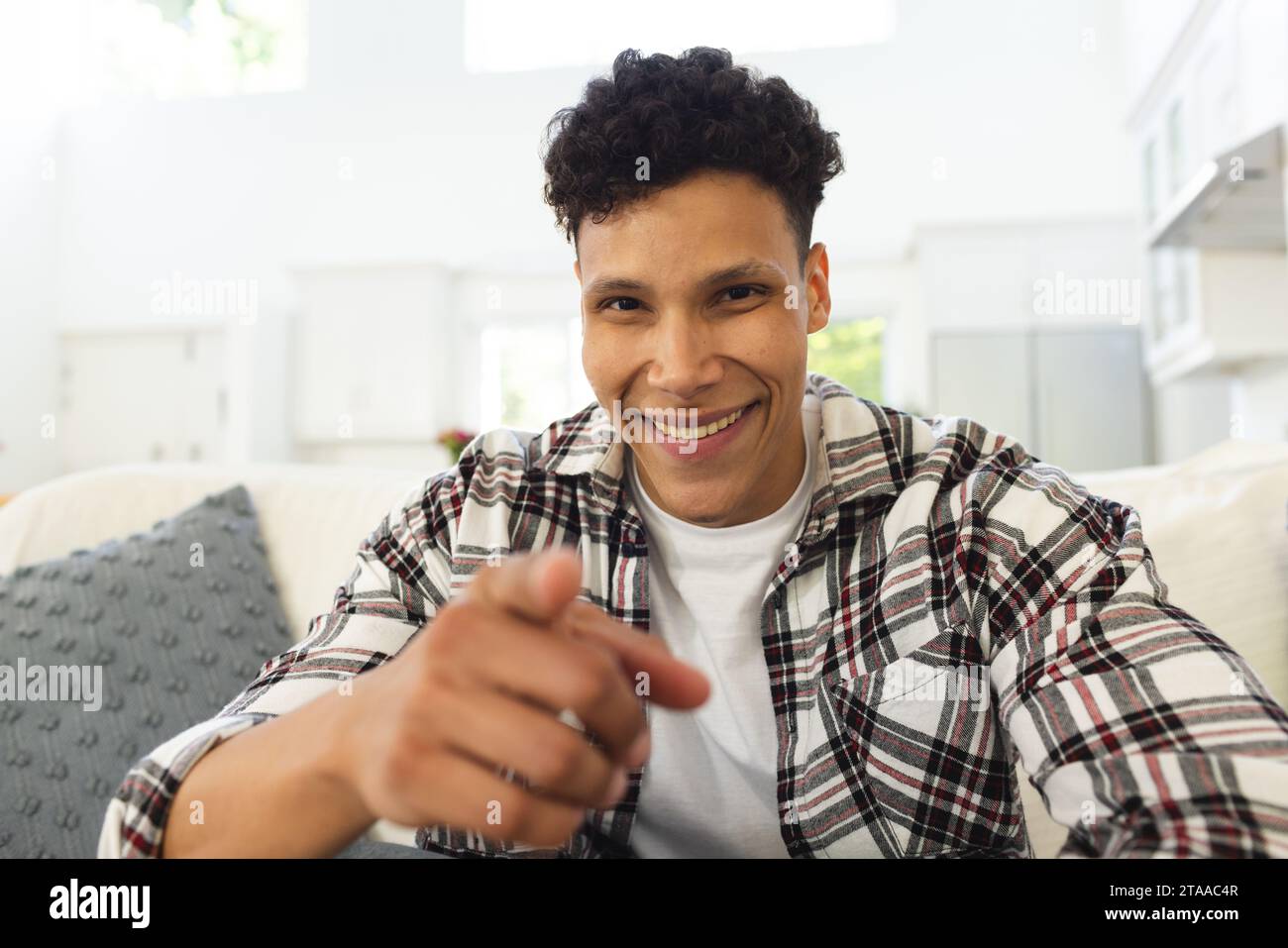 Ritratto di un felice uomo birazziale seduto sul divano e puntato con il dito a casa, spazio per copiare. Espressione, tempo libero, gesticolazione e vita domestica Foto Stock