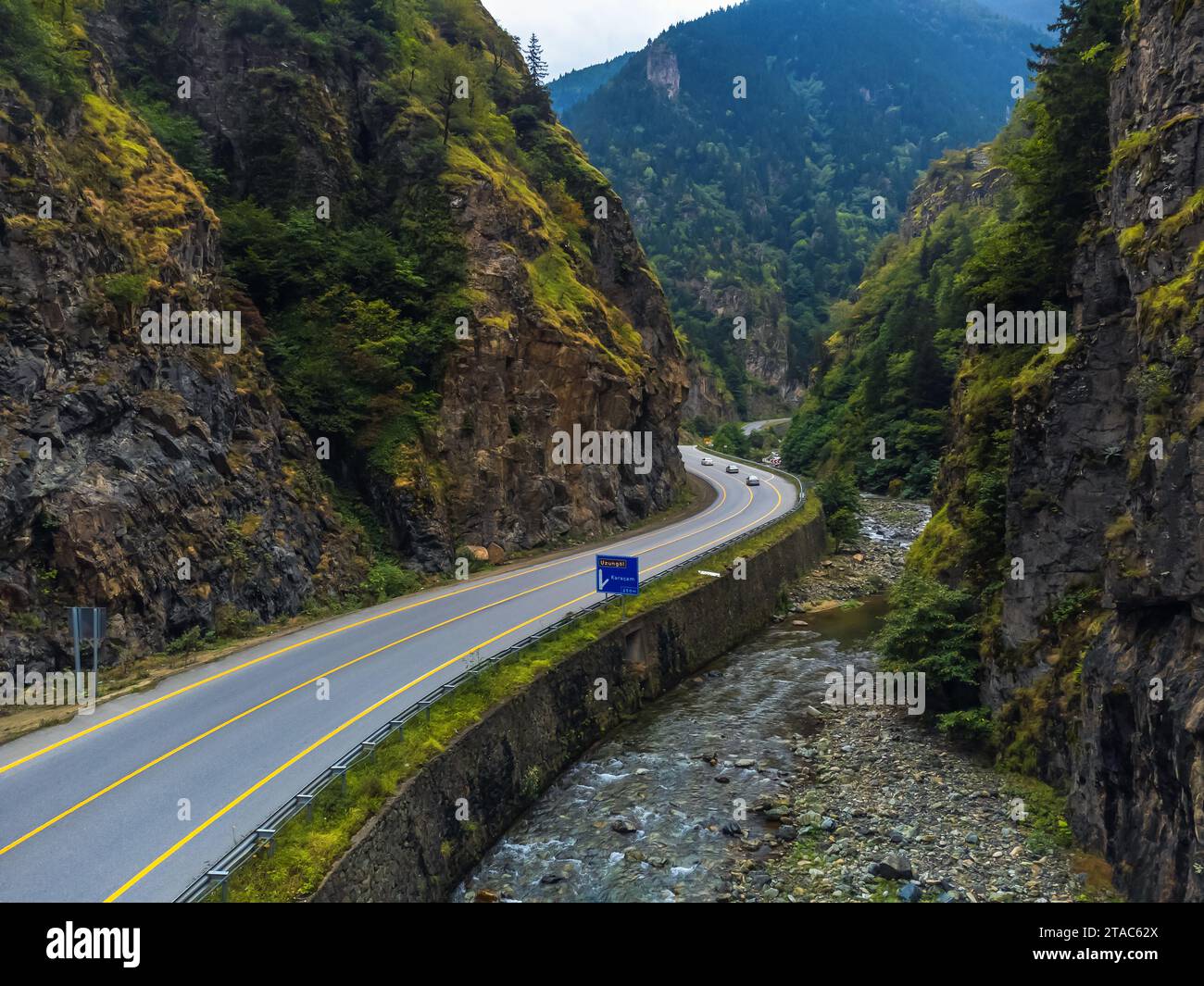 Bella strada asfaltata con un cartello stradale e auto in auto tra alte montagne rocciose di colore verde-marrone e un fiume verso Uzungel, Turchia, vista droni. Foto Stock